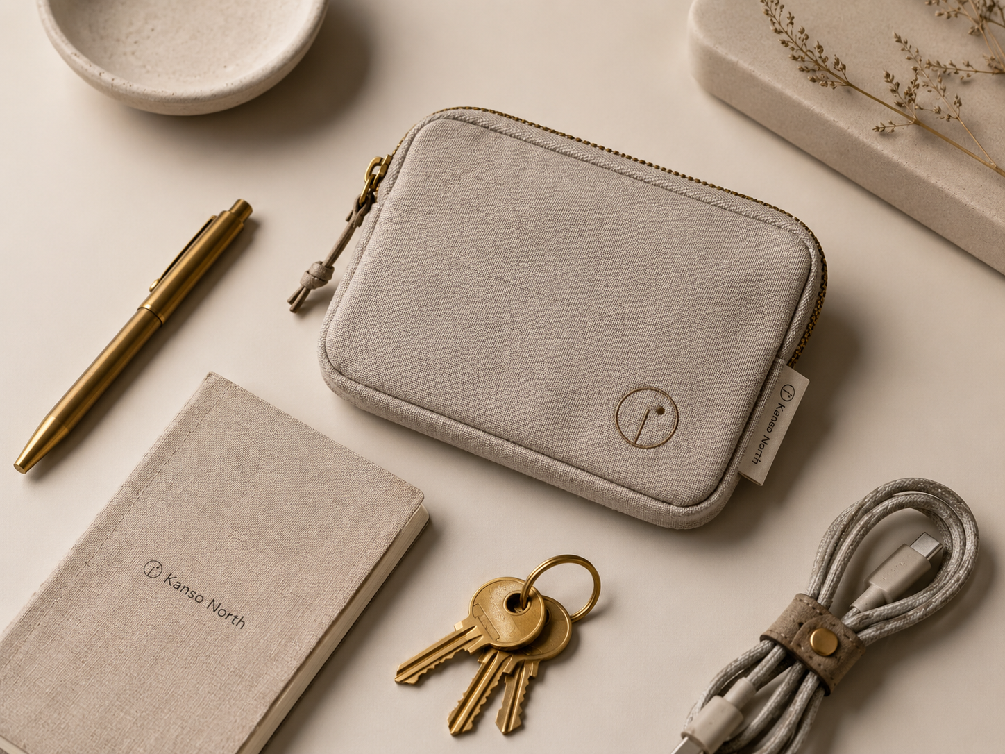 A linen notebook, brass pen, grey pouch and keys on a warm off-white surface